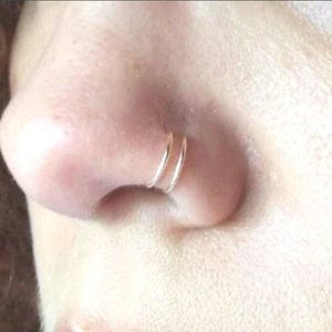 Op de afbeelding: Close-up van iemands neus met een dubbele neusring. De ring is van goud en wordt in het linker neusgat gedragen.