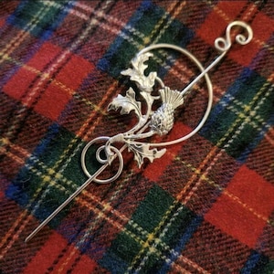 Può includere: Una forcina per capelli in metallo color argento con una cornice circolare e un motivo centrale a forma di cardo, caratterizzato da foglie dettagliate e un fiore testurizzato. Include un'asta lunga e dritta con un'estremità decorativa arricciata.