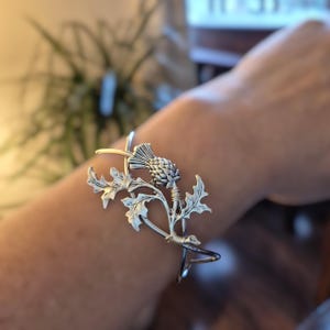 May include: Silver cuff bracelet with a detailed thistle design. The thistle, with its spiky head and leafy foliage, is the focal point. The bracelet wraps around the wrist, showcasing the intricate floral motif. The bracelet is made of metal.