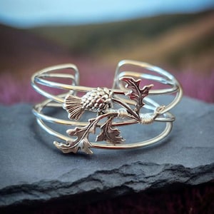 Op de afbeelding: Zilverkleurige manchetarmband met een gedetailleerd distelontwerp. De open armband heeft meerdere banden en een centraal distelmotief met bladeren. De sieraden worden getoond op een donkergrijze ondergrond met een vage paarse en groene achtergrond.