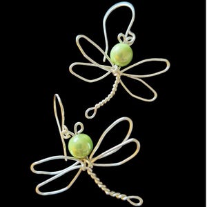 May include: A pair of dragonfly earrings. Each earring has a silver-coloured wire dragonfly design with delicate wings and a twisted body. A light green bead forms the dragonfly's head. The earrings hang from silver-coloured ear wires.