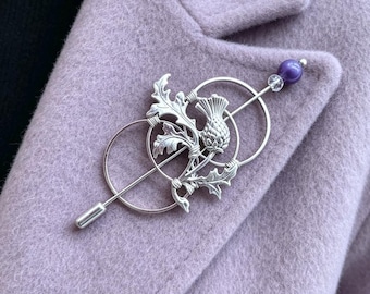May include: A silver-toned metal brooch featuring a detailed thistle design intertwined with two circular wire rings. The pin is topped with a small clear bead and a larger purple bead, secured horizontally across the textured fabric surface.