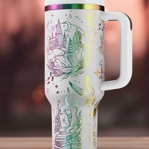 Könnte beinhalten: Weißer isolierter Becher mit Henkel, verziert mit einem farbenfrohen Design eines Schlosses, Büchern und anderen Illustrationen. Der Deckel und der Griff sind weiß, mit einem Regenbogenband am oberen Rand. Inklusive Strohhalm.