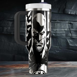May include: A stainless steel travel mug featuring a black and white illustration of Batman. The mug has a black handle, a white lid, and a straw. The design includes the Batman logo and bat silhouettes.