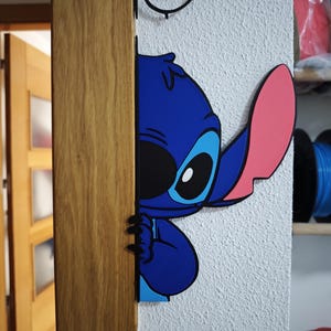 Può includere: Un personaggio dei cartoni animati blu e nero, con un orecchio rosa, che sbircia dietro un telaio di una porta in legno. Il personaggio ha grandi occhi blu ed è un personaggio popolare di un film Disney.