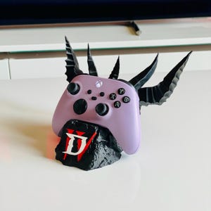 May include: A lavender Xbox controller with black accents, resting on a black base with a red and white "D" logo. The controller is adorned with black, stylized horns. The design is likely inspired by the video game Diablo.