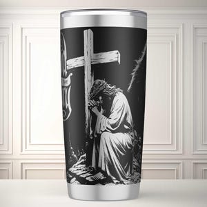 Może przedstawiać: Kubek termiczny ze stali nierdzewnej z czarno-białą ilustracją Jezusa modlącego się przed drewnianym krzyżem. Projekt zawiera tekst "I AM THE WAY". Kubek ma srebrną obręcz i podstawę. Temat religijny.