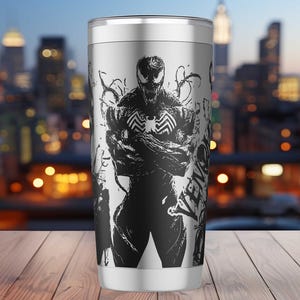 May include: A stainless steel tumbler featuring a black and white graphic of the character Venom. The design includes the character's face, a spider symbol, and the word "Venom". The tumbler is cylindrical and has a silver lid.