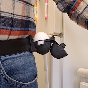 Peut inclure: Un clip de ceinture imprimé en 3D noir avec un œuf blanc à l'intérieur. Le clip est conçu pour maintenir un œuf en toute sécurité et peut être attaché à une ceinture.