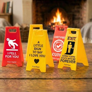 May include: A collection of red and yellow folding signs with humorous messages. The signs include phrases like "I Fell For You," "A Little Sign To Say I Love You," and "Sorry You're Stuck With Me Forever." The signs are on a wooden surface.