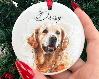May include: A round, white ceramic Christmas ornament featuring a watercolor portrait of a Golden Retriever dog. The dog's name, "Daisy", is written in cursive above the image. A red ribbon is attached for hanging.