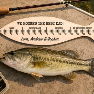 May include: A light-coloured wooden fish-shaped ruler with black text reading WE HOOKED THE BEST DAD! and Love, Andrew & Sophia. The ruler features a scale from 1 to 17 inches with labels including Bait, Throw Back, Keeper, An Awesome Story, and A Lie.