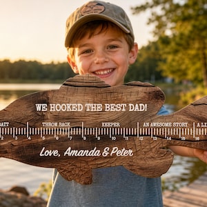 May include: A fish-shaped wooden plaque with a dark wood grain texture, featuring a ruler from 1 to 18 inches. Text reads: WE HOOKED THE BEST DAD! BAIT, THROW BACK, KEEPER, AN AWESOME STORY, A LIE, and Love, Amanda & Peter in white lettering.