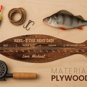 May include: A wooden fish-shaped ruler with a 17-inch scale, engraved with the text REEL-Y THE BEST DAD!, BAIT, THROW BACK, KEEPER, AN AWESOME STORY, A LIE, and Love, Micheal. Includes a fishing reel, a bobber, a coil of rope, a hook, and a fish.