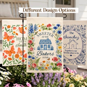 May include: Three decorative garden flags with different designs. One features orange butterflies and flowers with the text "Home of the Smith." Another has a house surrounded by flowers and the text "Sweeter at the Bakers Home." The third flag displays a house and the text "The Sanchez, Together is our favorite place to be."
