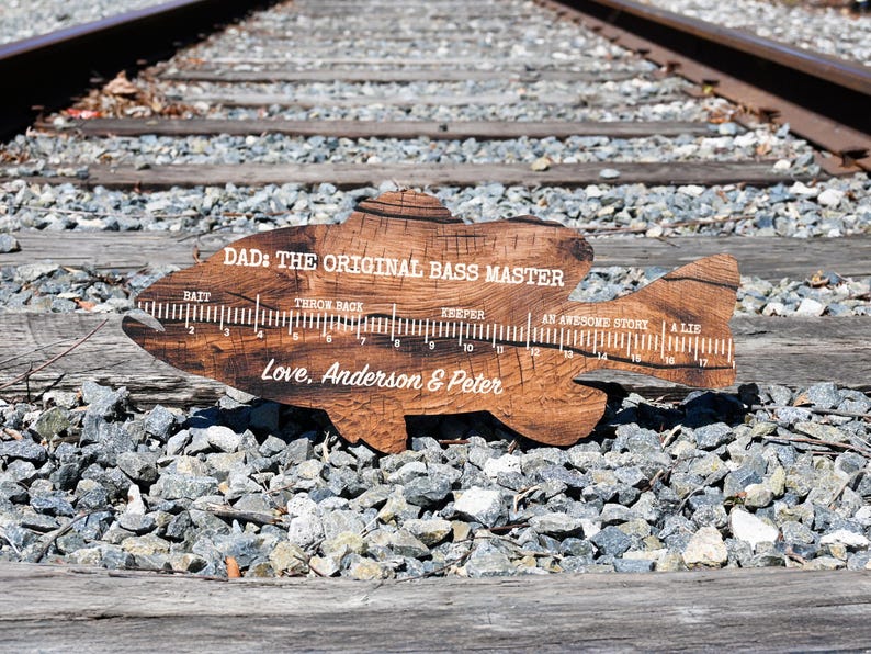 May include: A fish-shaped wooden board with a dark brown wood grain finish, featuring a white printed ruler from 1 to 17 inches. The text reads: DAD: THE ORIGINAL BASS MASTER, BAIT, THROW BACK, KEEPER, AN AWESOME STORY, A LIE, and Love, Anderson & Peter.