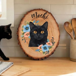 May include: A wooden slice featuring a black cat portrait with orange eyes, surrounded by blue and peach flowers. The name "Mochi" is written above the cat. A black cat is in the background, and wooden kitchen utensils are in a ceramic container.