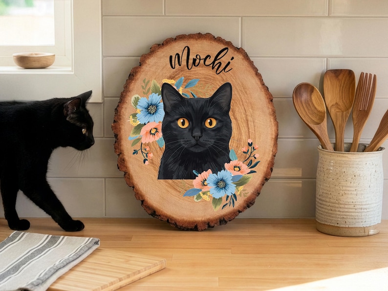 May include: A wooden slice featuring a black cat portrait with orange eyes, surrounded by blue and peach flowers. The name "Mochi" is written above the cat. A black cat is in the background, and wooden kitchen utensils are in a ceramic container.