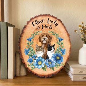 May include: A wooden plaque with a natural edge features a portrait of a dog and two cats, surrounded by blue floral accents. The names "Oliver, Lucky & Mochi" are written in script at the top. The plaque is displayed on a wooden surface.