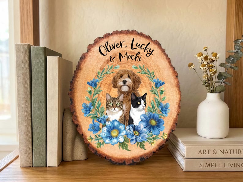 May include: A wooden plaque with a natural edge features a portrait of a dog and two cats, surrounded by blue floral accents. The names "Oliver, Lucky & Mochi" are written in script at the top. The plaque is displayed on a wooden surface.