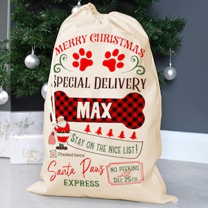 K&ouml;nnte beinhalten: Ein beiger Weihnachtsgeschenksack mit dem Text "Merry Christmas" und "Special Delivery". Er zeigt rote Pfotenabdr&uuml;cke, ein knochenf&ouml;rmiges Design mit einem rot-schwarzen Karomuster und den Namen "MAX". Die Tasche enth&auml;lt auch eine Illustration von Santa Claus und den Ausdruck "Santa Paws Express".