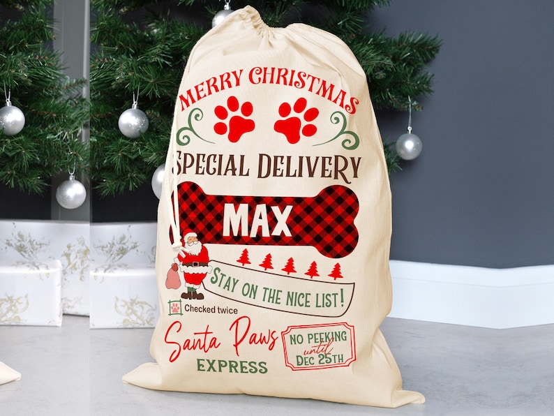 K&ouml;nnte beinhalten: Ein beiger Weihnachtsgeschenksack mit dem Text "Merry Christmas" und "Special Delivery". Er zeigt rote Pfotenabdr&uuml;cke, ein knochenf&ouml;rmiges Design mit einem rot-schwarzen Karomuster und den Namen "MAX". Die Tasche enth&auml;lt auch eine Illustration von Santa Claus und den Ausdruck "Santa Paws Express".