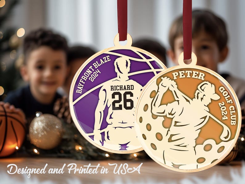 May include: Two gold-coloured Christmas ornaments featuring a basketball player and a golfer. The basketball ornament is purple with the text "BAYFRONT BLAZE 2024" and "RICHARD 26". The golf ornament is orange with the text "PETER GOLF CLUB 2024".
