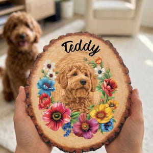 May include: A wooden slice featuring a portrait of a dog named Teddy, surrounded by a floral wreath. The dog is a light brown color, and the flowers are in shades of red, blue, yellow, and purple. The name "Teddy" is written in black script.