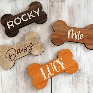 May include: Four wooden dog bone-shaped name tags. The tags are in various shades of brown and orange, with names like "Rocky", "Milo", "Daisy", and "Lucy" in white script. The tags are on a light wood surface.