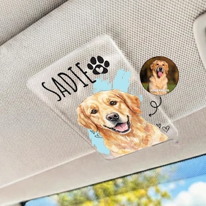 May include: A clear, rectangular car visor clip featuring a Golden Retriever illustration. The clip has the name "Sadie" in black script, a paw print, and a small photo of the dog. Decorative hearts and swirls are also present.