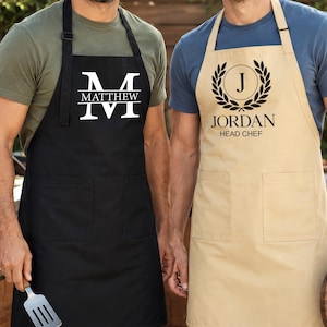 May include: Two aprons, one black and one tan, are displayed. The black apron has the name "Matthew" and the letter "M" in white. The tan apron has the name "Jordan" and the words "Head Chef" with a laurel wreath design.
