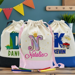 May include: Three personalized drawstring bags with different designs. The first bag is black and white with a dinosaur and the name "Dani". The second bag is pink and white with a crown and the name "Natalie". The third bag is blue and white with a monster truck and the name "Kevin".