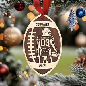 May include: A brown wooden football bauble with a gold outline. The bauble features a football player in a black uniform with the number "03" on the back. The text "COUGARS" and "JOHNSON" are also on the bauble, along with the year "2024".