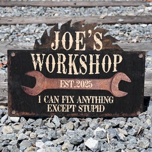 Könnte beinhalten: Ein rustikales Holzschild für eine Werkstatt mit der Aufschrift "JOE'S WORKSHOP" über einer Schraubenschlüssel-Grafik und dem Satz "I CAN FIX ANYTHING EXCEPT STUPID". Das Schild befindet sich auf einem Hintergrund aus Bahngleisen und Schotter.