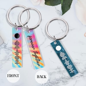 May include: Two silver keychains with a rectangular tag. The front of the tag has a rainbow tie-dye pattern with a black circle and a barcode. The back of the tag has a rainbow tie-dye pattern with the text "forever together". The other keychain has a blue marble pattern with a white circle and a barcode.