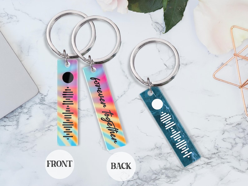 May include: Two silver keychains with a rectangular tag. The front of the tag has a rainbow tie-dye pattern with a black circle and a barcode. The back of the tag has a rainbow tie-dye pattern with the text "forever together". The other keychain has a blue marble pattern with a white circle and a barcode.