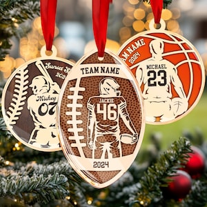 May include: Three gold-coloured Christmas baubles featuring a baseball, football, and basketball. Each bauble has a player silhouette and a personalised name, number, and year. The baseball bauble says "Mickey 07 Sunset Sr 2022", the football bauble says "TEAM NAME Jackie 46 2024", and the basketball bauble says "TEAM NAME Michael 23 2024".