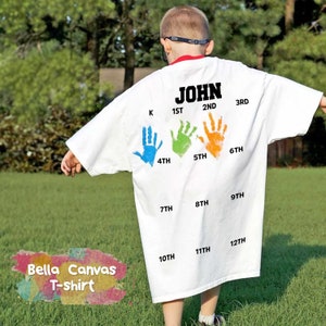 May include: White t-shirt with the name "JOHN" printed on the back. The shirt has handprints in different colors representing each grade from kindergarten to 12th grade.