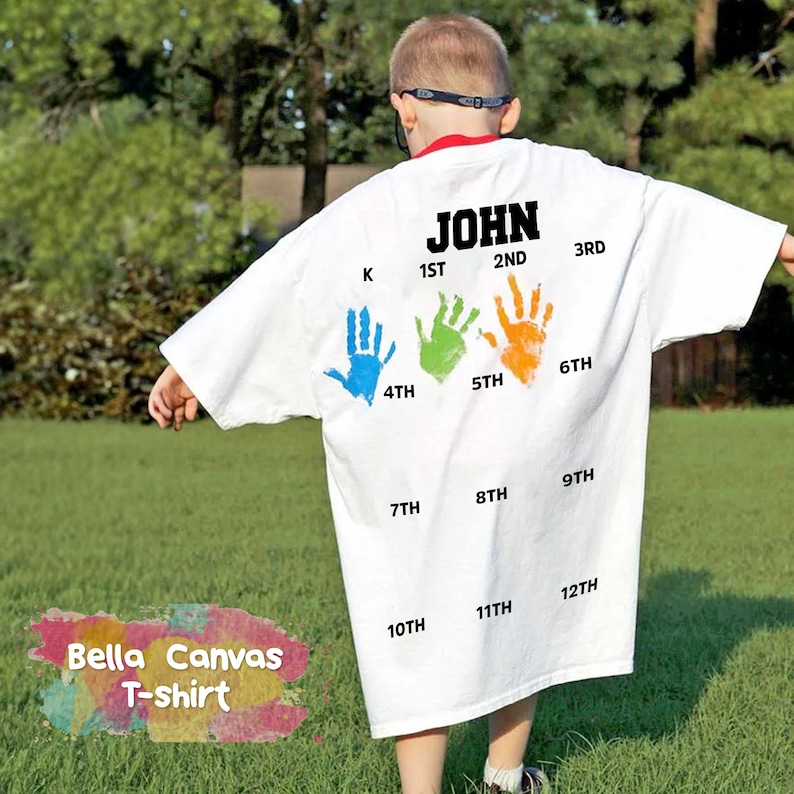 May include: White t-shirt with the name "JOHN" printed on the back. The shirt has handprints in different colors representing each grade from kindergarten to 12th grade.