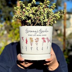 May include: A white ceramic planter with the text "Granny's Garden" and "Make Life More Grand" in black script. The planter features colorful floral illustrations and is filled with a green plant.