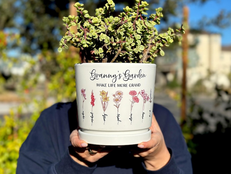 May include: A white ceramic planter with the text "Granny's Garden" and "Make Life More Grand" in black script. The planter features colorful floral illustrations and is filled with a green plant.
