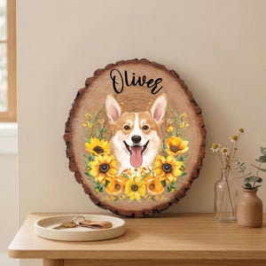 May include: A wooden plaque with a corgi illustration surrounded by sunflowers. The corgi has a happy expression with its tongue out. The name "Oliver" is written above the corgi in black script.