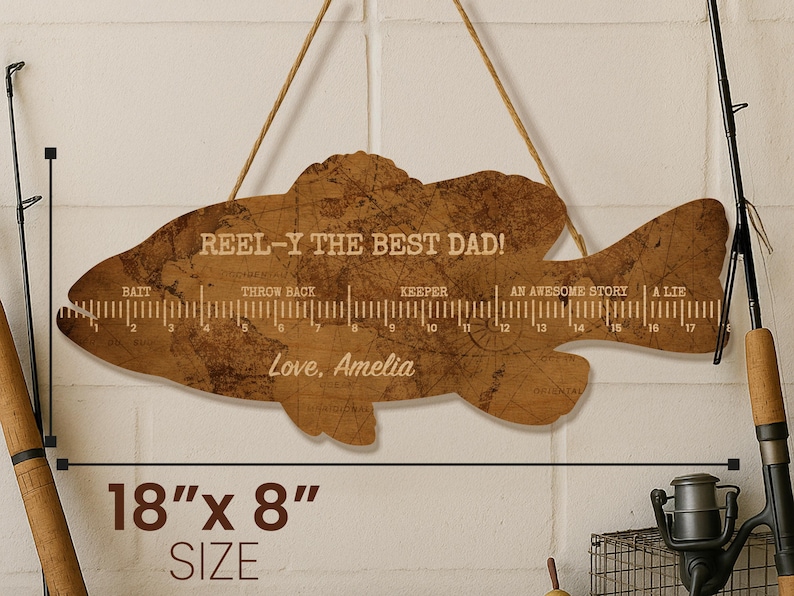 May include: A fish-shaped wooden wall hanging with a brown map-like texture, featuring a ruler from 0 to 17 inches, the text REEL-Y THE BEST DAD!, labels for Bait, Throw Back, Keeper, An Awesome Story, A Lie, and Love, Amelia, measuring 45.7 by 20.3 cm.