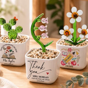 May include: Three decorative glass plant sculptures in white square pots. One is a green cactus, another has pink flowers, and the third has white and yellow flowers. Each pot has a message and a name.