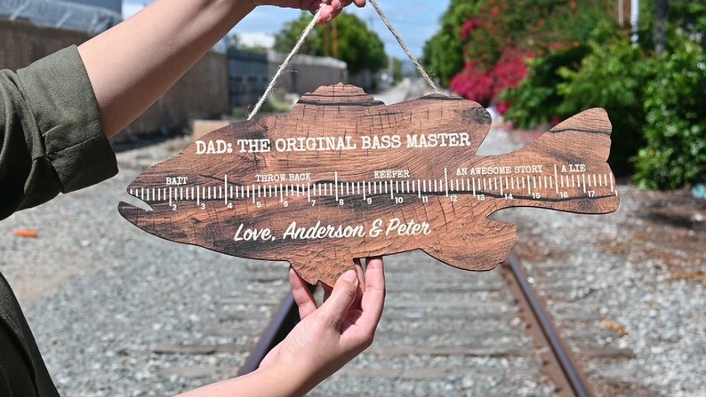 May include: A fish-shaped wooden wall hanging with a dark wood grain finish. It features a printed ruler from 0 to 17 inches with labels: Bait, Throw Back, Keeper, An Awesome Story, A Lie. Text reads: DAD: THE ORIGINAL BASS MASTER and Love, Anderson & Peter.