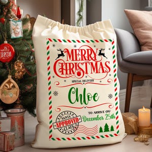 K&ouml;nnte beinhalten: Ein gro&szlig;er, cremefarbener Weihnachtssack mit Kordelzug. Er zeigt "Merry Christmas" in Rot, den Namen "Chloe" in Gr&uuml;n und das Datum "25. Dezember". Das Design umfasst Rentiere, Schneeflocken und ein "Santa Approved" Siegel. Ein festlicher Geschenksack.