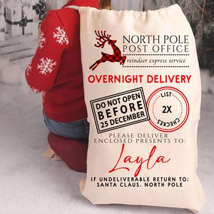 K&ouml;nnte beinhalten: Ein gro&szlig;er, cremefarbener Weihnachtsgeschenksack mit dem Text "North Pole Post Office" und "Overnight Delivery". Ein rotes Rentier-Motiv und der Name "Layla" sind ebenfalls sichtbar. Der Sack ist mit Geschenken gef&uuml;llt.