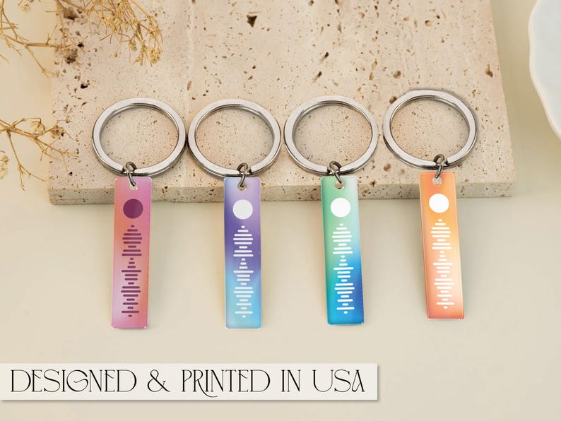 May include: Four silver keychains with colorful gradient rectangular tags. Each tag features a white circle and a white sound wave design. The keychains are arranged in a row on a light beige surface.