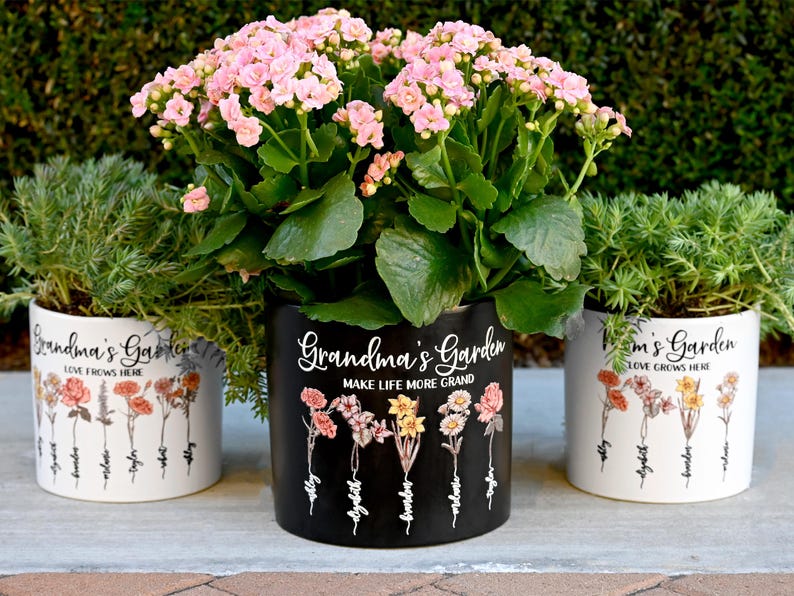 May include: Three ceramic flower pots with plants. The center pot is black with the text "Grandma's Garden MAKE LIFE MORE GRAND". The other two pots are white with the text "Grandma's Garden LOVE GROWS HERE". Each pot has floral illustrations.
