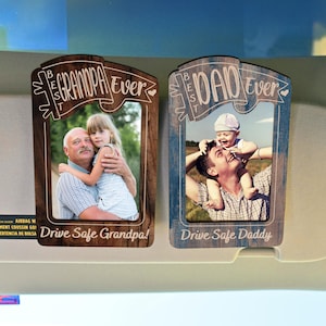 May include: Two wooden car visor photo frames. One frame says "Best Grandpa Ever" and the other says "Best Dad Ever". Each frame has a photo of a person with a child. The bottom of each frame says "Drive Safe".